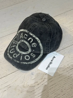 Svart keps från Acne Studios - Cool svart keps från Acne Studios med tvättad look och stort vitt tryck framtill. Justerbar rem baktill för perfekt passform. Perfekt accessoar för en avslappnad och trendig stil.