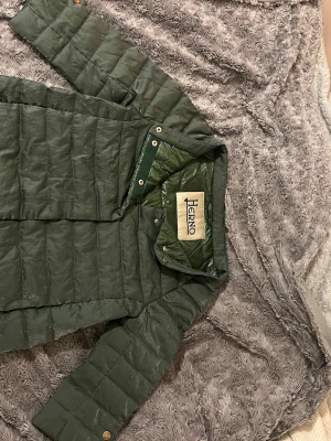 Grön dunjacka från Herno - Stilren grön dunjacka från Herno som är 3/4 ärmad med diskret glansig yta och raka sömmar. Jackan har tryckknappar framtill och en enkel, kraglös design. 