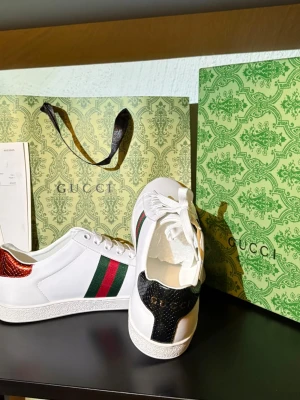 Gucci Ace vita sneakers - Gucci Ace sneakers i vitt läder med klassiska gröna och röda. Snygg och tidlös modell med vita skosnören och präglad sula. Perfekta för dig som vill ha en lyxig touch till din outfit.
