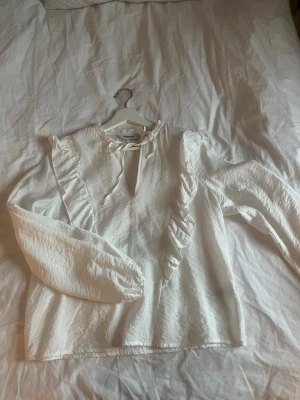 Vit volangblus från Kappahl - Supersöt vit blus från Kappahl med volangdetaljer längs framsidan och knytning vid halsen. Blusen har långa ärmar med resår vid ärmslut och är tillverkad i ett tunt tyg 💗