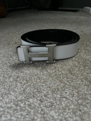 Vitt skärp med silverfärgat H-spänne - Snyggt vitt bälte med ett stort H-format spänne i silverfärg. Bältet har en stilren och minimalistisk design med svarta kanter och passar perfekt till jeans eller byxor. Materialet ser ut att vara skinn eller liknande.