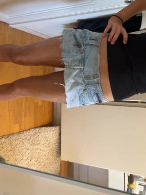 Ljusblå denimshorts från RIVARS - Ljusblå fenimkjol från RIVARS med låg midja, råa kanter och flera detaljer som knappar och dragkedjor framtill. Shortsen har en unik design med slitningar. Köpte i london. 