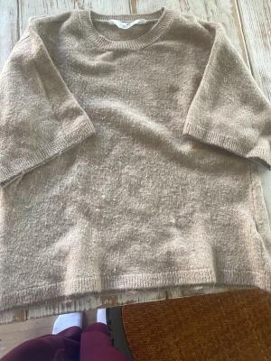 Beige stickad tröja med kortarmad längd  - Mysig beige stickad tröja med rund halsringning och kortarmad. Tröjan har en avslappnad passform och ribbstickade detaljer vid ärmslut och nederkant. Perfekt för lager på lager och enkel att matcha med olika stilar.