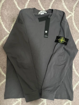 Grå långärmad topp Stone Island - Säljer en grå långärmad topp från Stone Island med klassisk rund hals och ikonisk patch på ärmen. Tillverkad i mjuk bomull som känns skön mot huden. Perfekt för dig som gillar stilrena och tidlösa plagg med streetwear-vibe.