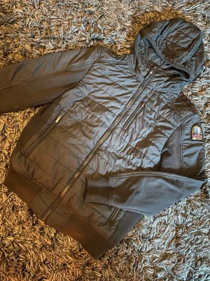 Svart pufferjacka från Parajumpers - Snygg svart pufferjacka från Parajumpers med huva och dragkedja framtill. Jackan har quiltad framsida och ärm, samt ribbade muddar och en patch med logga på ärmen. Perfekt för kalla vinterdagar och riktigt streetig look.