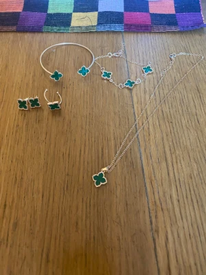Smyckesset med gröna fyrklöver - Säljer ett smyckesset med halsband, armband, örhängen och ring i silverfärg med gröna fyrklöverformade hängen. Setet har en modern och stilren design som sticker ut med sina klara gröna detaljer och fina former.