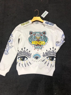 Vit Kenzo hoodie med tigertryck - Snygg vit hoodie från Kenzo med färgglatt tigertryck och ögonmotiv framtill. Kenzo-logga på axlarna och detaljerade mönster på ärmarna. Perfekt statement-plagg för dig som vill sticka ut med din stil.