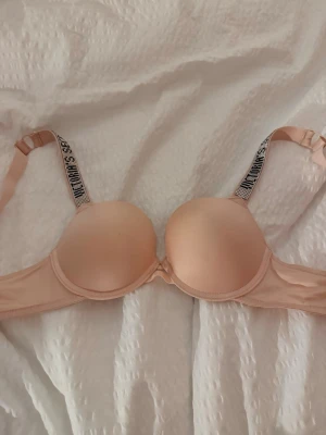 Beige push-up bh från Victoria's Secret - Säljer en beige push-up bh från Victoria's Secret med vadderade kupor och svarta axelband med logotyp. Bh:n har bygel och justerbara axelband för bästa passform. Materialet är mjukt och bekvämt, perfekt under ljusa kläder.
