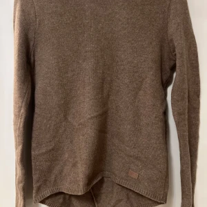 Brun stickad tröja-Holebrook - Säljer en brun stickad tröja med lång ärm och unik asymmetrisk nederkant. Tröjan har en enkel design utan synliga mönster eller detaljer och passar perfekt till höst och vinter. Mjuk och skön att bära.