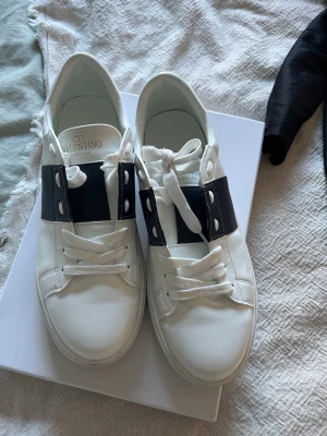 Valentino Garavani vita sneakers - Snygga vita sneakers från Valentino Garavani med bred blå läderdetalj över snörningen och klassiska vita skosnören. Skorna har en låg profil och stilren design som passar perfekt till streetwear och trendiga outfits.. obs på kartong står stl 45 men är felmärkta, passar ca 42,5 Jag har köpt dessa på Plick men de var fel storlek så kan ej svara på äkthet etc. Ser oanvända ut 