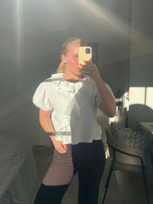 Vit blus med volang och puffärm🩵 - Trendig vit blus med stora volanger längs halsringningen och puffiga korta ärmar. Blusen har en lätt strukturerad yta och rak passform, perfekt för en stilren och modern look. Materialet ser ut att vara bomull eller en bomullsblandning.