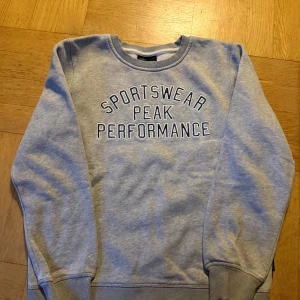 Grå sweatshirt Peak Performance - Cool grå sweatshirt från Peak Performance med texten 'SPORTSWEAR PEAK PERFORMANCE' på bröstet. Tröjan har rund halsringning och ribbade muddar vid ärmslut och nederkant. Perfekt för en avslappnad look.