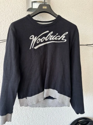 Svart Woolrich tröja med logga - Cool svart tröja från Woolrich med stor vit logotyp broderad på bröstet. Tröjan har ribbade muddar och nederkant i ljusgrått, rund halsringning och långa ärmar. Perfekt för dig som gillar klassisk streetstyle och vill ha något bekvämt.