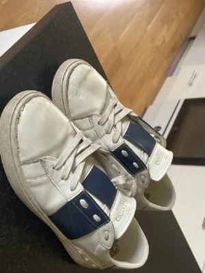 Valentino vita sneakers med blå detalj - Snygga vita sneakers från Valentino med bred blå läderdetalj och silvriga nitar på sidan. Klassisk låg modell med vita skosnören och logga på plösen. Perfekta för dig som gillar stilrena men ändå unika skor. Se bilder för att bedöma skick, pris kan diskuteras