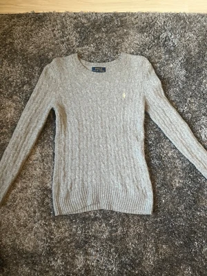 Grå kabelstickad tröja Polo Ralph Lauren - Säljer en grå kabelstickad tröja från Polo Ralph Lauren med rund hals och diskret broderad logga på bröstet. Tröjan har långa ärmar och ribbade muddar vid ärmslut och nederkant. Perfekt för dig som gillar klassisk och stilren design.