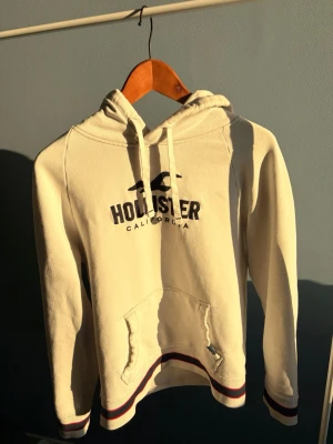 Vit hoodie från Hollister - Vit hoodie från Hollister med svart logga och text på bröstet. Klassisk känguruficka framtill och huva med dragsko. Muddar och nederkant har rödblåa ränder som ger en sportig vibe. Perfekt för chill dagar och enkel att matcha med jeans eller joggers. Vintage🙌🏼