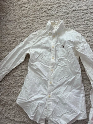 Vit skjorta från Polo Ralph Lauren - Klassisk vit skjorta från Polo Ralph Lauren med broderad logga på bröstet. Skjortan har lång ärm, knappar hela vägen och en snygg krage. Perfekt för dig som gillar stilrena plagg och ikoniska märken.