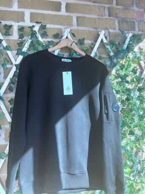 Svart sweatshirt från C.P. Company - Cool svart sweatshirt från C.P. Company med rund hals och en ikonisk linsdetalj på ärmen. Tröjan har en enkel och stilren design med ribbade muddar och är tillverkad i mjukt material som känns skönt mot huden.