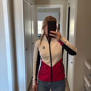 Träningsjacka från dählie i vitt och rosa - Tränings jacka från dählie i vitt, svart och rosa med dragkedja framtill och hög krage. Jackan har långa ärmar och sportig design med logga på bröstet. Perfekt för löpning eller annan träning utomhus.