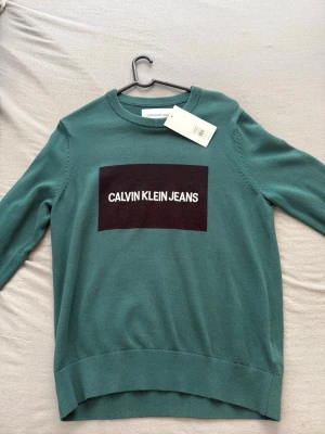Grön Calvin Klein Jeans tröja - Stilren grön tröja från Calvin Klein Jeans med svart patch och vit logga framtill. Tröjan har rund halsringning och ribbade muddar vid ärmslut och nederkant. Perfekt för en avslappnad och trendig look. Oanvänd!