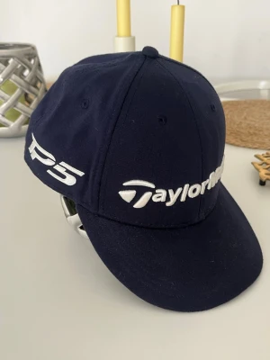Marinblå TaylorMade keps (helt ny!) - Snygg mörkblå keps från TaylorMade med böjd skärm och vit broderad logga framtill och STEALTH texten på sidan. Helt ny aldrig använd. Klassisk design med ventilationshål och justerbar baksida. Perfekt för golfbanan!