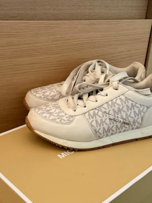 Vita Michael Kors sneakers med logga - Snygga vita sneakers från Michael Kors med klassiskt MK-mönster i grått och beige. Skorna har vita snören, gummisula och detaljer i läderimitation samt en metallisk logga på sidan. Perfekta för dig som gillar stilrena och trendiga skor.