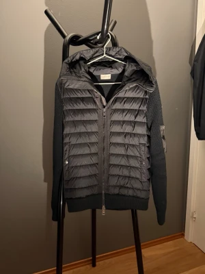Svart cardigan från Moncler - Snygg svart cardigan från Moncler med stickade ärmar och vadderad front. Jackan har huva, dragkedja framtill och Moncler-logga på ärmen. Perfekt för kalla dagar och riktigt trendig stil.