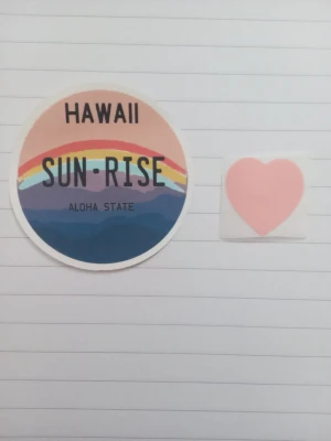 Klistermärken  - Två klistermärken: ett runt med texten 'HAWAII SUN-RISE ALOHA STATE' i pastellfärger med regnbåge, samt en med ett ljusrosa hjärta. Säljer båda tillsammans för 20 kr.