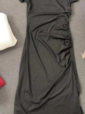 Svart långklänning från SHEIN - Säljer en svart långklänning från SHEIN i storlek S. Klänningen har korta ärmar och snygg rynkning på sidan som ger en cool detalj. Tillverkad i mjukt och stretchigt syntetmaterial som sitter skönt på kroppen.