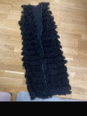 Lång svart pälsväst med fransar - Snygg lång svart pälsväst med fransiga detaljer i flera lager. Västen har en rak passform och är fodrad på insidan för extra komfort. Perfekt för att lyfta din outfit med en trendig touch. Materialet är fuskpäls.