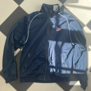 Blå Nike track jacket med vita detaljer - Snygg blå track jacket från Nike med dragkedja, vita kontrastlinjer och broderad logga på bröstet. Jackan har två fickor med dragkedja och ribbade muddar vid ärmslut och nederkant. Vintage och trendig! Perfekt nu till sommar och vår! 