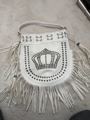 Vit handväska med fransar och nitar (krona) - Säljer min vita handväska i skinnimitation med långa fransar och silverfärgade nitar i ett kronmönster på framsidan. Ass snygg väska! 4 mindre hål på backsidan av väskan! Kommer oxå med ett cross body band!