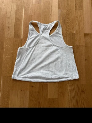 Vit croppad linnetopp från Adidas - Säljer en vit croppad linnetopp från Adidas med stort logotryck framtill. Toppen har brottarrygg och är tillverkad i mjuk bomull, perfekt för träning eller chill. Enkel och sportig design som är lätt att matcha.