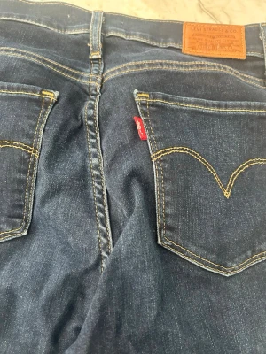 Mörkblå Levi's jeans med kontrastsömmar - Säljer ett par klassiska mörkblå jeans från Levi's med tydliga kontrastsömmar och ikoniska bakfickor med gul söm. Jeansen har en skön passform och är tillverkade i slitstark denim. Perfekta för dig som gillar tidlös stil och vill ha ett par jeans som alltid funkar.