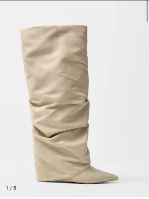 Beige slouchy stövel med spetsig tå - Trendig beige stövel med slouchy design och spetsig tå. Stöveln har en hög, rynkad skaft och kilklack som ger en cool siluett. Tillverkad i mockaimitation för en mjuk och lyxig känsla. Perfekt för dig som vill sticka ut med din stil. Nytt skick 