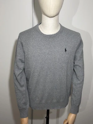 Grå stickad tröja från Ralph Lauren - Snygg grå stickad tröja från Ralph Lauren med rund hals och diskret broderad logga på bröstet. Tröjan har långa ärmar och ribbade muddar vid ärmslut och nederkant. Perfekt för dig som gillar stilren och enkel design.