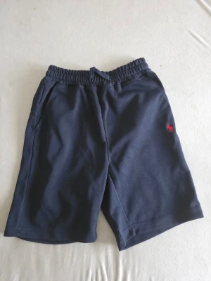 Marinblå shorts från Polo Ralph Lauren - Snygga marinblå shorts från Polo Ralph Lauren med elastisk midja och fickor på sidorna. Klassisk röd logga broderad på vänster ben. Perfekta för sommardagar och avslappnade tillfällen.