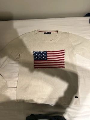 Vit stickad tröja med USA-flagga från GANT - Säljer en vit stickad tröja från GANT med ett stort broderat USA-flagga motiv på bröstet. Tröjan har rund halsringning och långa ärmar. Materialet är mjukt och bekvämt, perfekt för att ge en cool och avslappnad look.