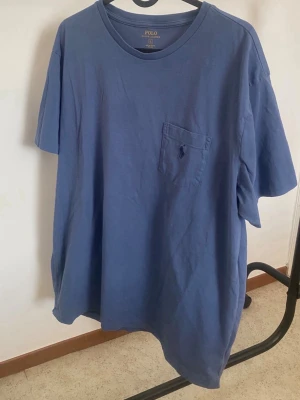 Blå t-shirt från Polo Ralph Lauren - Säljer en blå t-shirt från Polo Ralph Lauren med klassisk rund hals och bröstficka med broderad logga. T-shirten är i bomull och har en loose passform med korta ärmar. Perfekt för dig som gillar enkel och stilren design.