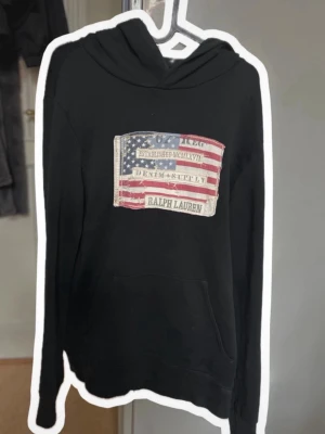 Denim and supply hoodie - Svart hoodie från Ralph Lauren med stor patch framtill som föreställer en amerikansk flagga och texten 'Denim & Supply'. Hoodien har huva och känguruficka. Materialet ser ut att vara mjukt bomullstyg, perfekt för en avslappnad och cool stil.