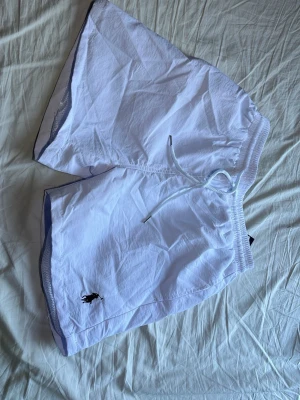 Vita shorts från Polo Ralph Lauren -  Går att diskutera priset. Snygga vita bad-shorts från Polo Ralph Lauren med klassisk broderad logga i svart på benet. Shortsen har elastisk midja med snörning och är tillverkade i ett lätt och bekvämt material, perfekta för varma dagar.