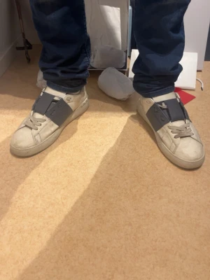 Vita sneakers med grå detalj - Säljer ett par vita sneakers från Valentino med bred grå rem över vristen och klassisk snörning. Skorna har en låg profil och är tillverkade i läder med subtila nitar på hälen. Perfekta för dig som gillar stilrena och exklusiva sneakers.