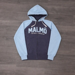 Malmö FF hoodie med huva - Blå och ljusblå hoodie från Malmö FF med dragkedja framtill och fickor. Stor text 'MALMÖ FOTBOLLFÖRENING' på bröstet. Klassisk huva med vita snören och raglanärmar. Perfekt för dig som vill representera ditt lag. Troligtvis i bomullsblandning.
