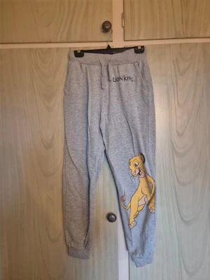 Grå Lion King mjukisbyxor - Säljer ett par grå mjukisbyxor med tryck från The Lion King. Byxorna har en cool Simba-illustration på benet och logga på höften. De har resår och snörning i midjan samt muddar vid bensluten. Perfekta för chill och avslappnade dagar.  Alla kläder rollas innan dom skickas hem då det finns katt i hemmet🐈 Jag stryker och tvättar INTE innan jag skickar det får du stå för själv så det blir som du vill ha det🤗