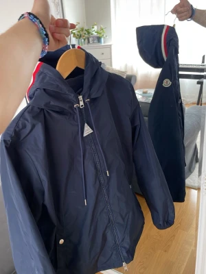 Mörkblå Moncler vindjacka med huva - Snygg mörkblå vindjacka från Moncler med huva och dragkedja. Jackan har vita och röda detaljer på huvan samt Moncler-logga på ärmen. Materialet är lätt och vattenavvisande, perfekt för blåsiga dagar. Jackan har fickor med tryckknappar och justerbar dragsko vid huvan. Storlek M men den passar för folk som har S också