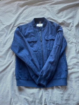 Blå bomberjacka från dressman - Snygg blå bomberjacka från dressman med dragkedja framtill, två bröstfickor med lock och ribbade muddar. Insidan har ett unikt blått mönster med naturmotiv. Tillverkad i ett mjukt tyg som känns skönt mot huden.