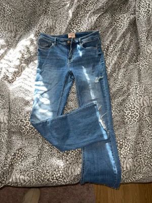 Blå slitna jeans från Gina Tricot - Säljer ett par blå jeans från Gina Tricot med raka ben och slitningar på låret och knät. Jeansen har klassisk femficksmodell, knappgylf och är tillverkade i bomull med lite stretch. Perfekta för en avslappnad och trendig look.