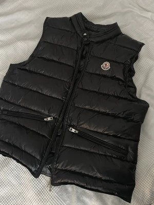 Svart dunväst  Moncler - Snygg svart dunväst från Moncler med glansig finish och klassisk logga på bröstet. Västen har två dragkedjeförsedda fickor framtill och hög krage. Perfekt för lager-på-lager och riktigt stilren till alla outfits. storlek 3 
