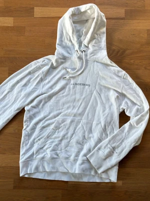 Vit hoodie från J.Lindeberg - Snygg vit hoodie från J.Lindeberg med klassisk huva. Framtill finns en stor magficka och diskret logga tryckt på bröstet. Tillverkad i mjukt bomullsmaterial som känns skönt mot huden. Perfekt för en avslappnad och stilren look.