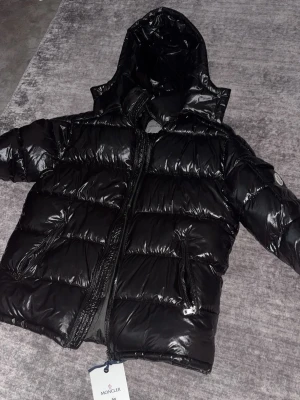 Svart dunjacka med huva från Moncler - Svart, glansig dunjacka från Moncler med huva och två dragkedjeförsedda fickor. Jackan har quiltad design och Moncler-logga på ärmen. Perfekt för kalla vinterdagar och har en modern, sportig look.